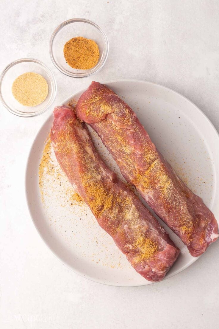 two pork tenderloins on a white plate with rub ingredients in glass bowls.