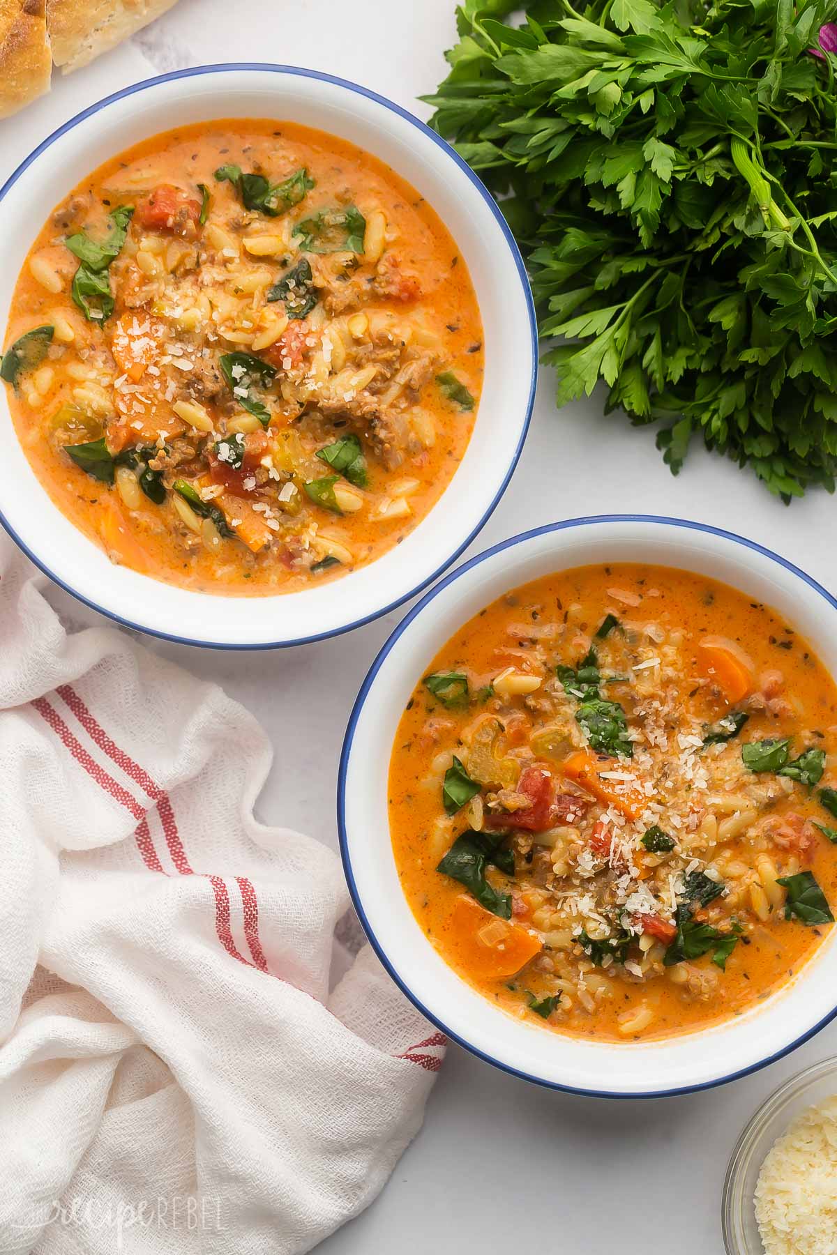 two full bowls of sausage orzo soup with shredded cheese and parsley around it.