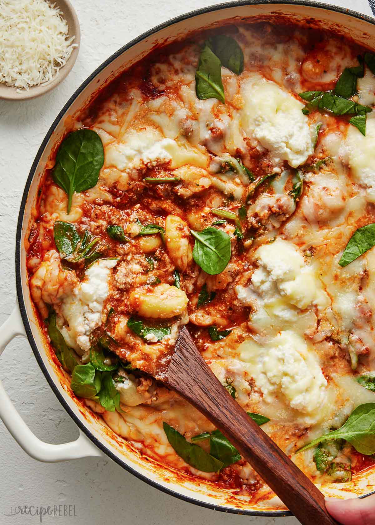 a wooden ladle in pot of gnocchi bake with shredded cheese beside.