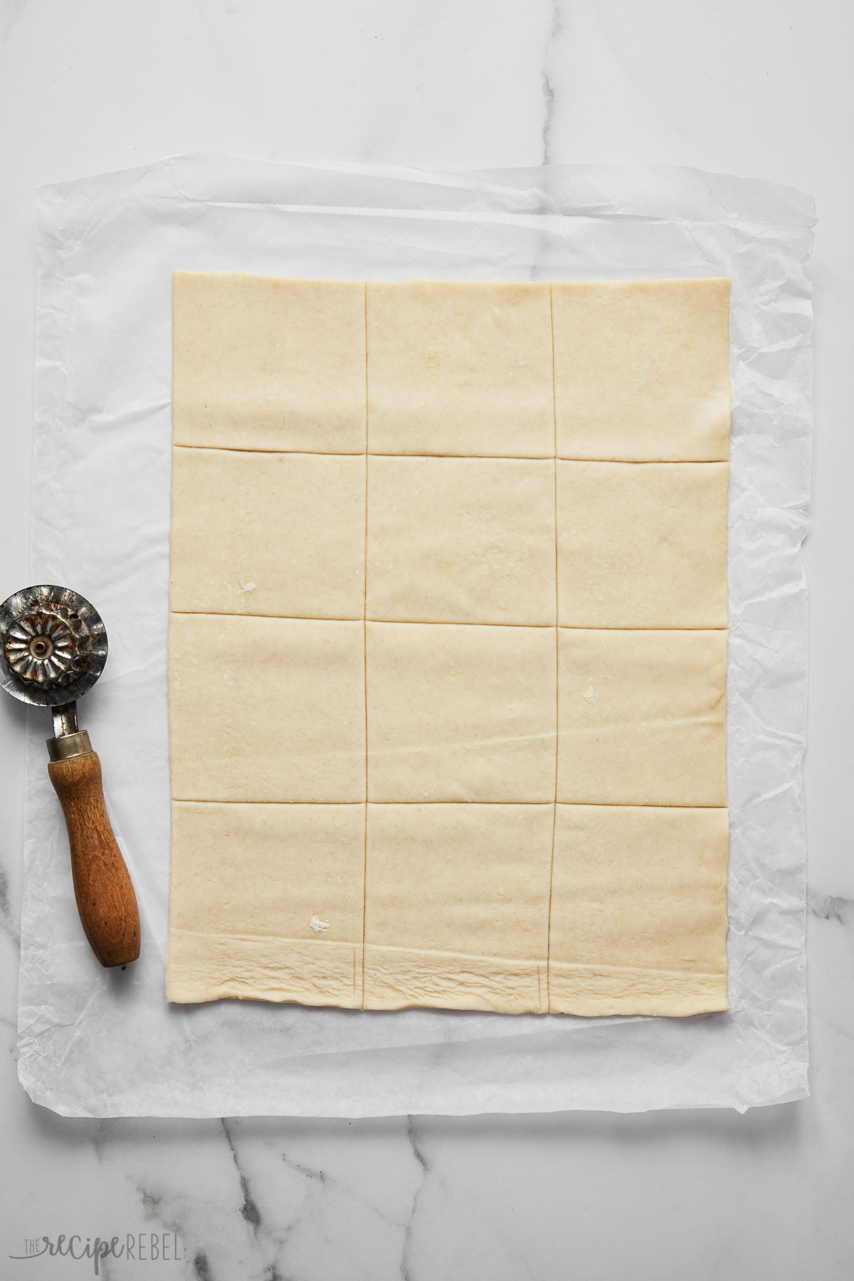 Top view of a sheet of puff pastry sliced into 12 squares on a white worktop with a pastry cutter next to it.