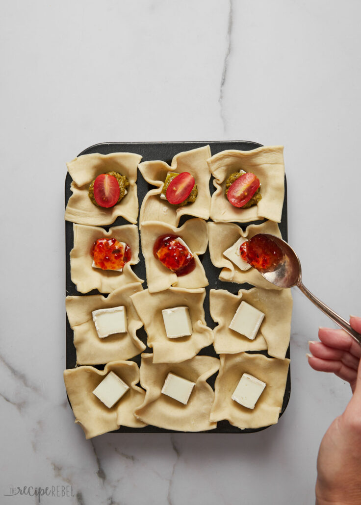 Top view of pastry squares in a mini muffin pan being filled with brie and tomato with a spoon.