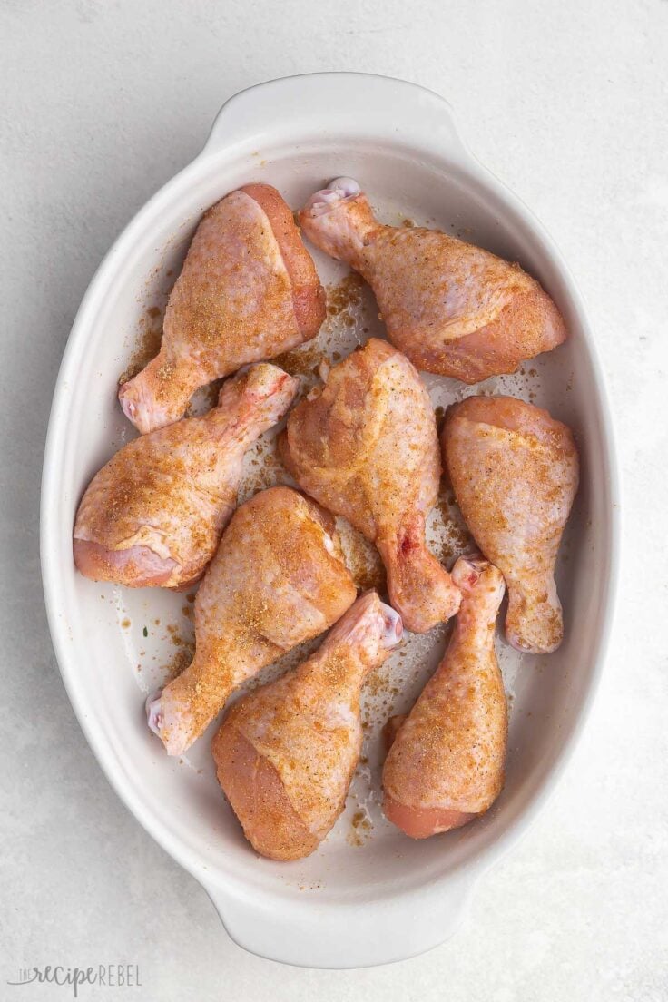 uncooked chicken drumsticks with seasoning in a white baking dish.
