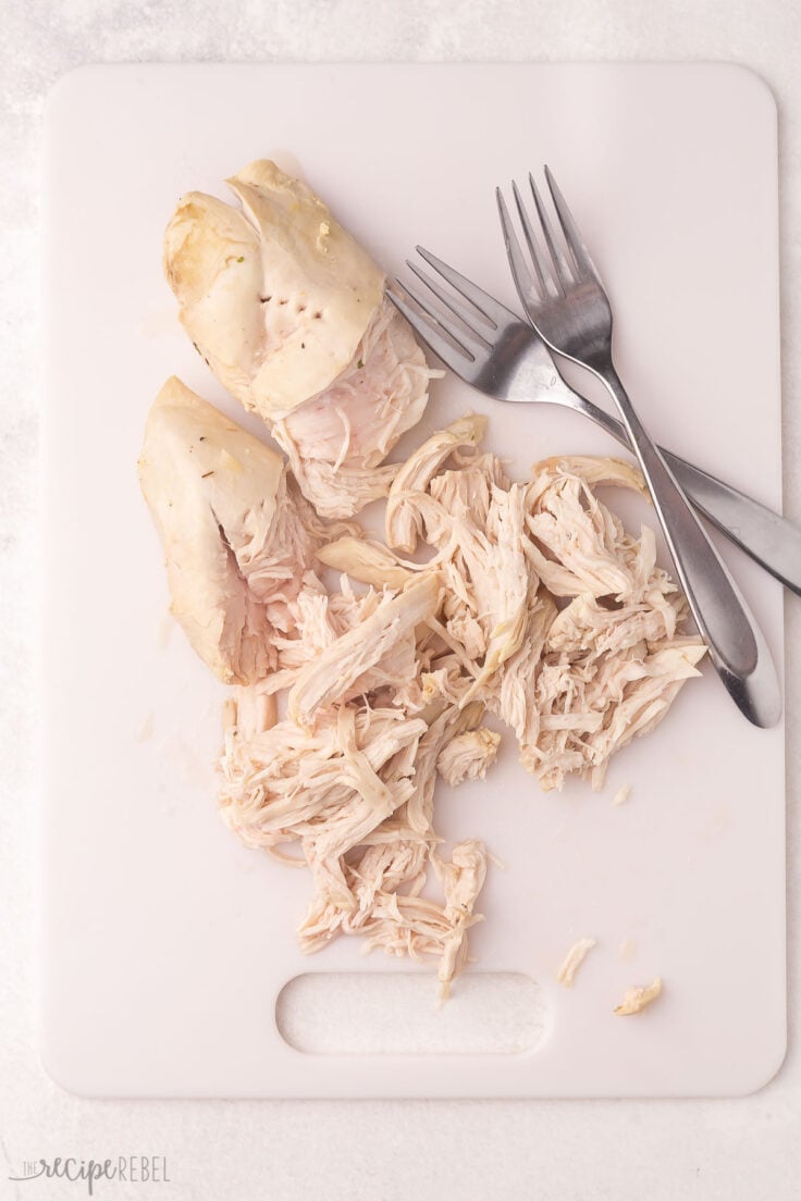 shredded chicken breast lying on a white cutting board beside two forks.