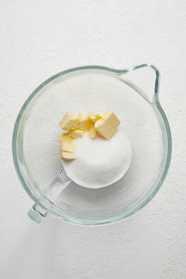 glass mixing bowl with cubed butter and sugar in it on white surface.