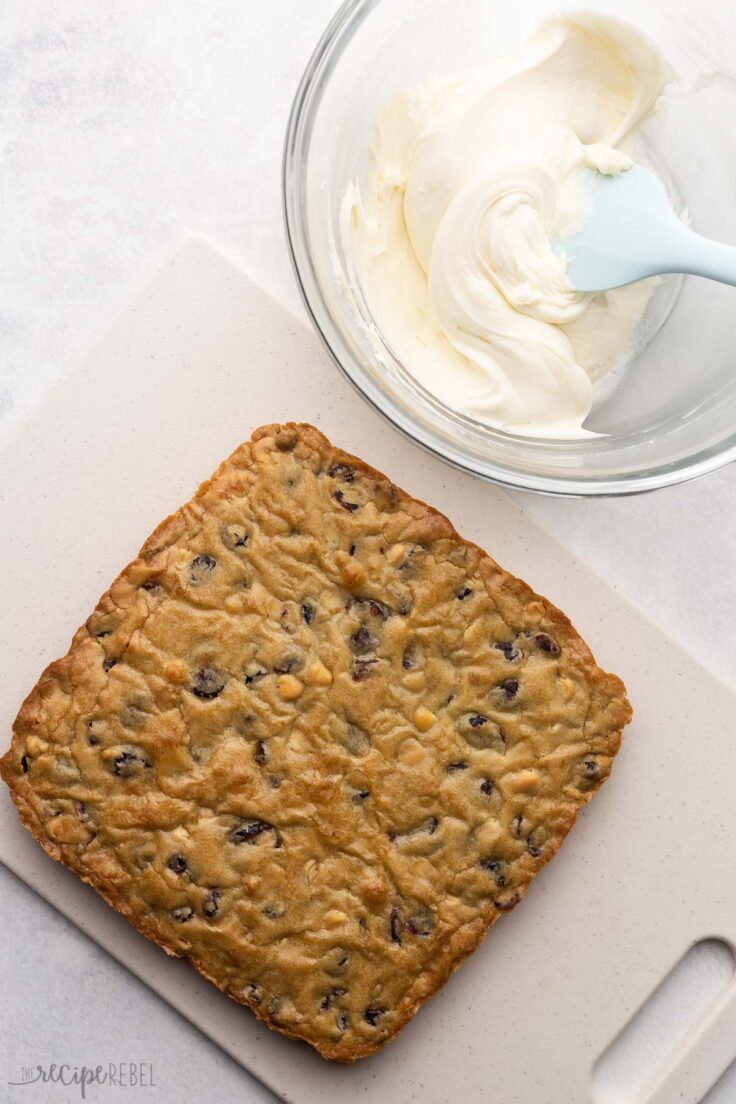 baked cranberry bliss bars on a cutting board with a bowl of icing beside.