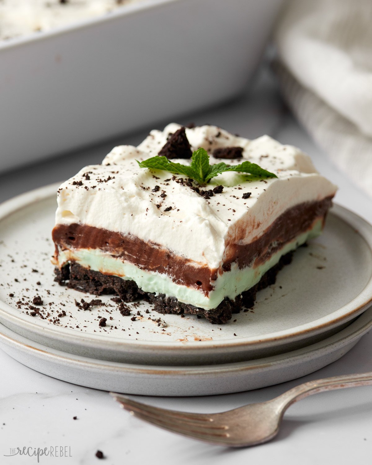 No Bake Chocolate Mintย Lasagna is the perfect dessert for the holidays! With an Oreo cookie crust, it's has layers of mint cream cheese and chocolate pudding, topped with homemade whipped cream, Andes mints, fresh mint leaves, and chocolate curls! #chocolate #dessert #christmas | christmas dessert | christmas baking | no bake dessert | chocolate lasagna | dessert lasagna | christmas recipes | easy dessert fork and grey plate with piece of chocolate mint lasagna on it.
