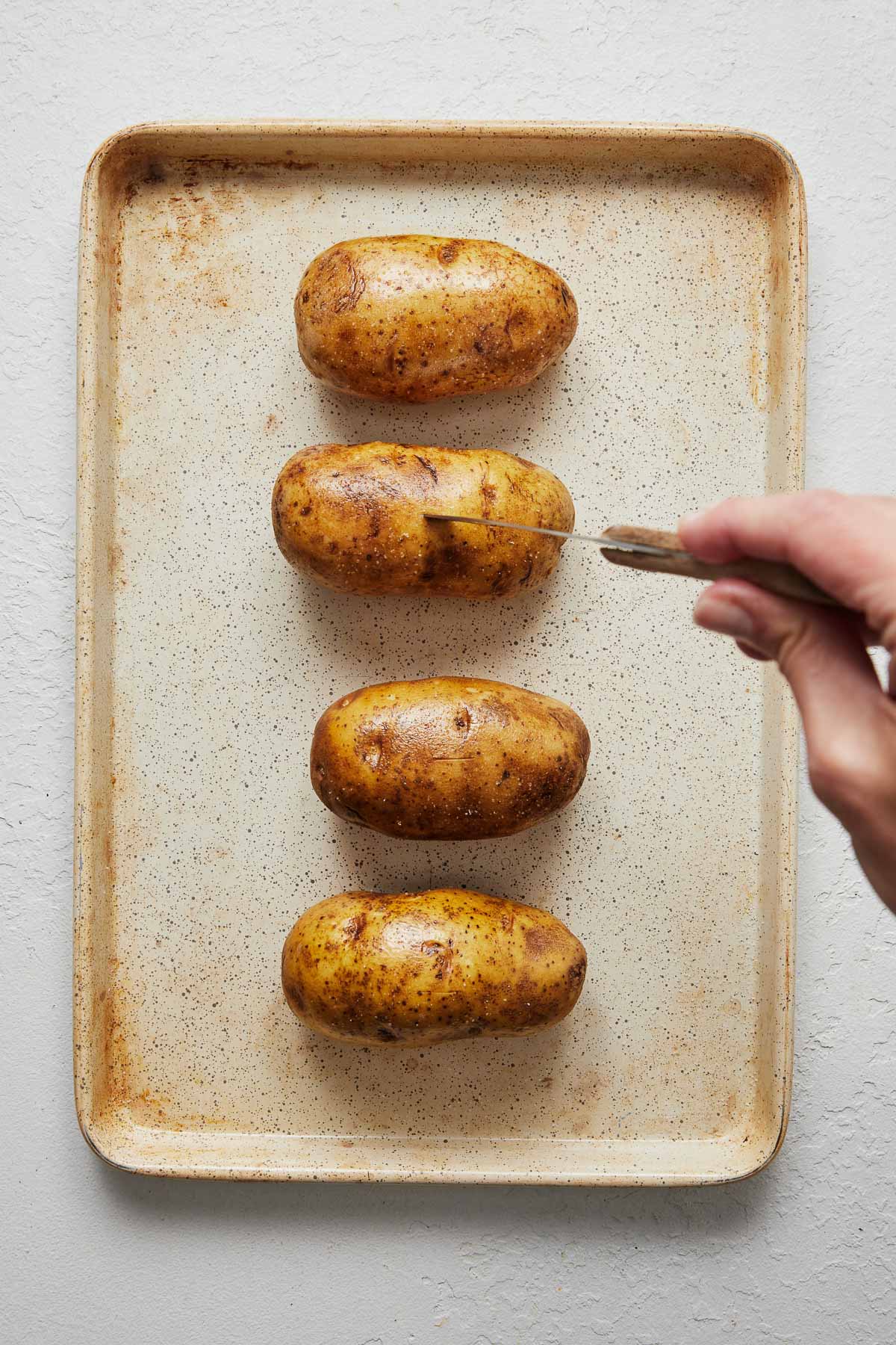 Top view of four potatoes on a chopping board, and a knife is making holes in them.