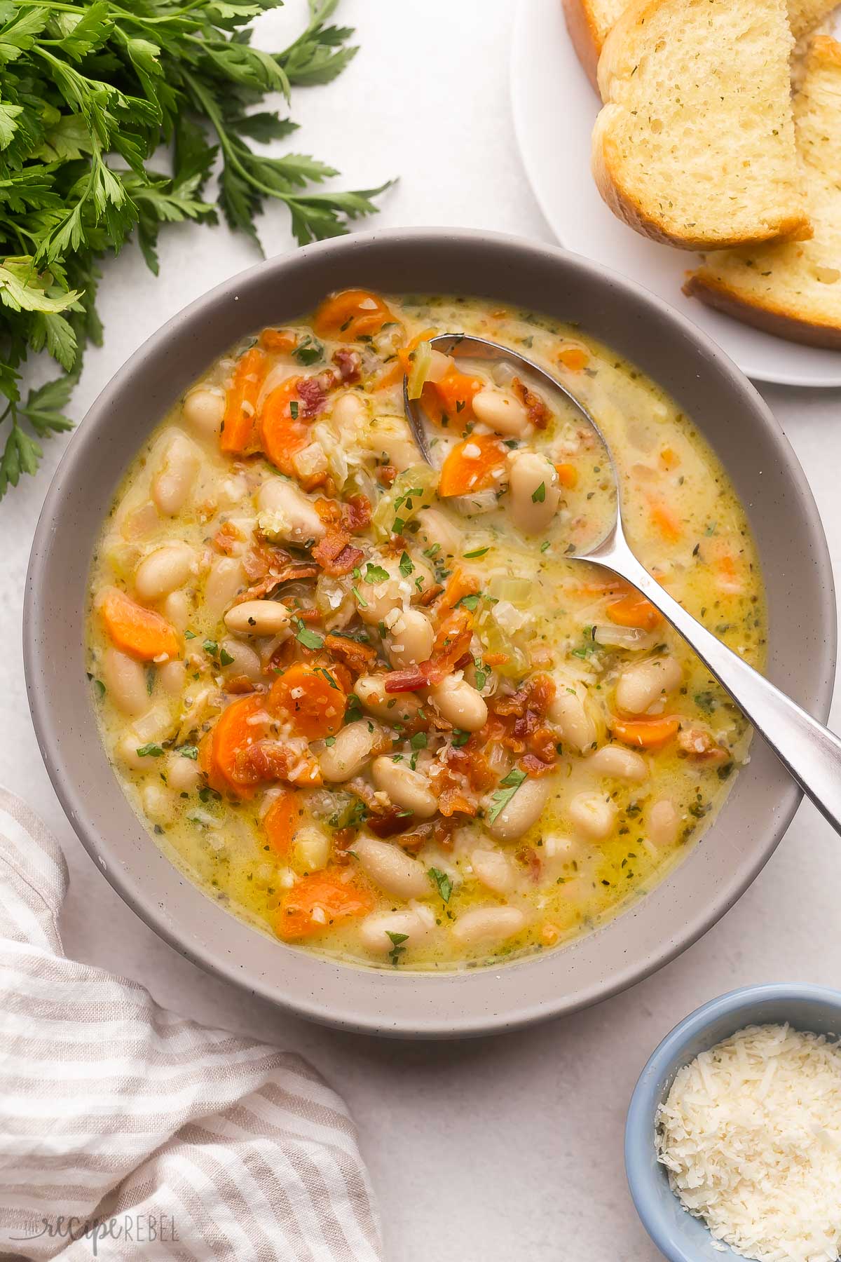 grey bowl filled with bacon white bean soup with parsley and grated cheese beside.