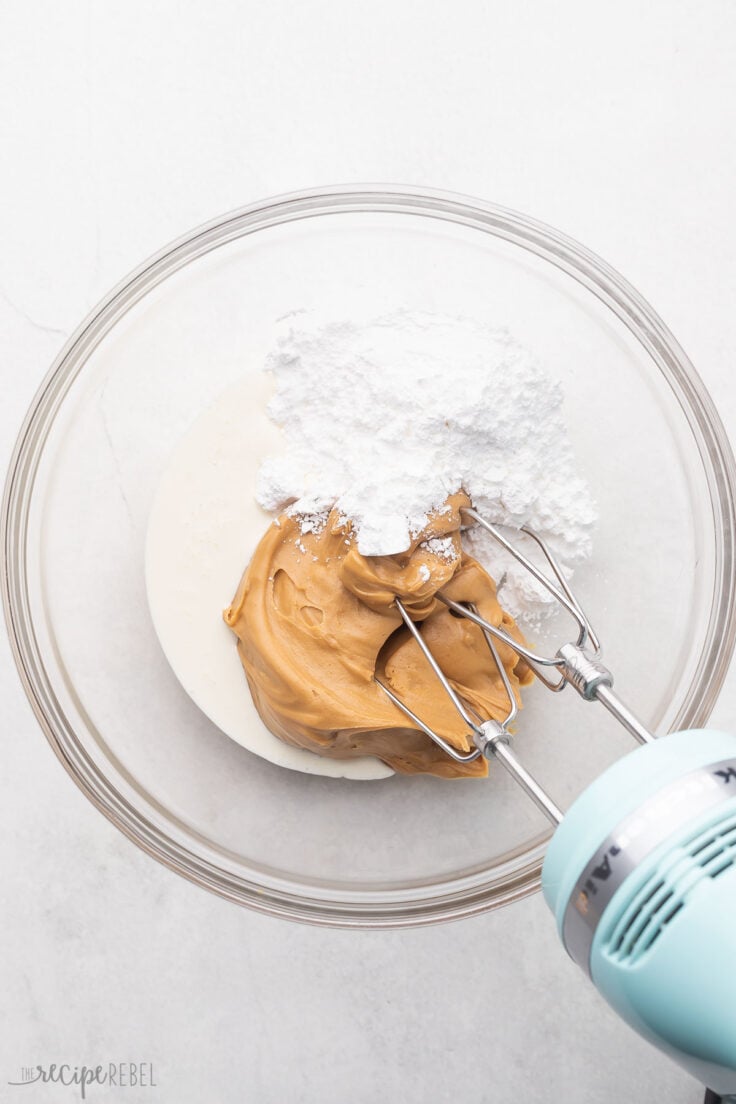 Top view of a glass mixing bowl with peanut butter, icing sugar, and cream being beaten with an electric mixer.