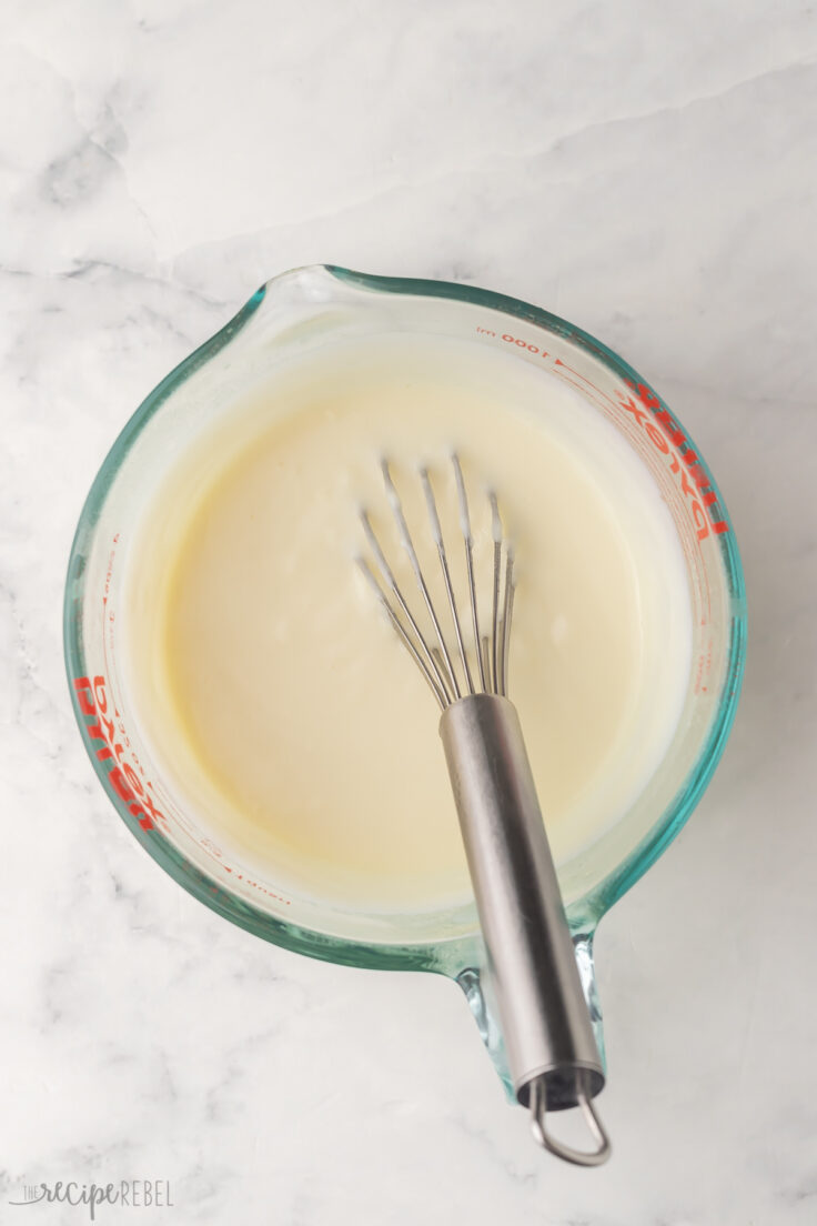 glass measuring cup filled with mixed white sauce and metal whisk.