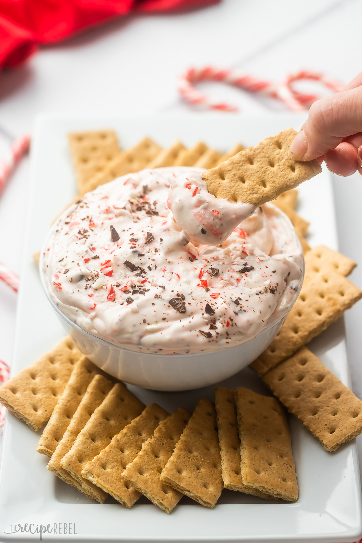 a hand holding a graham cracker scooping peppermint bark cheesecake dip out of bowl.
