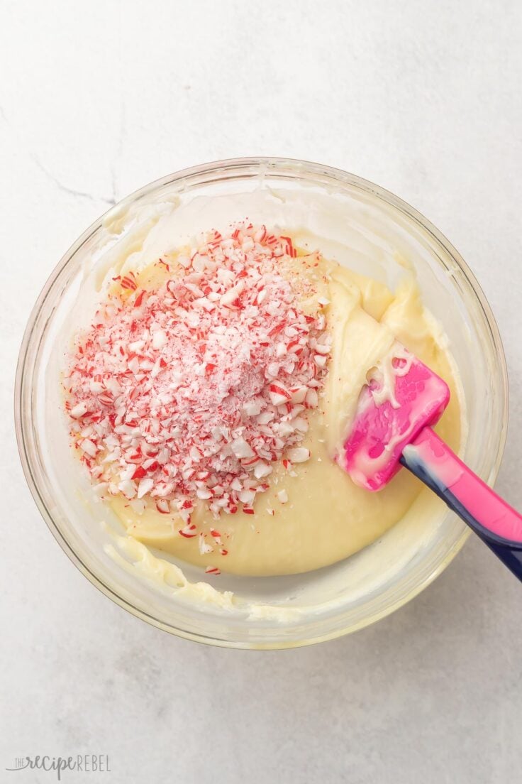 Top view of a glass mixing bowl with melted white chocolate and evaporated milk, with peppermint bark pieces on top.
