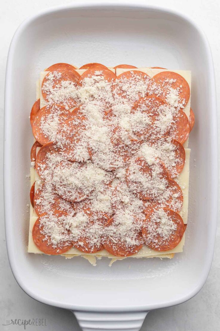 A layer of pepperoni sprinkled with parmesan cheese, on top of the cheese slices, on the pepperoni, on the slider rolls.