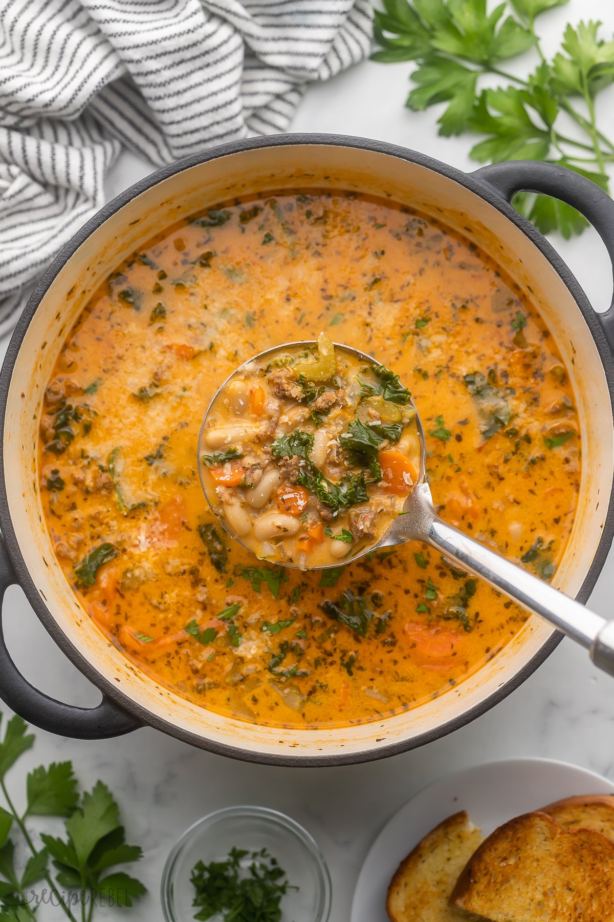 This Tuscan White Bean Soup is made with sautéed carrots, onion and celery plus white beans, Italian sausage and kale in a flavorful, creamy tomato-flavored broth. It is a cozy, hearty meal that's perfect with some crusty bread for dunking! #soup #dinner #recipe | dinner recipes | dinner ideas | bean soup | canned beans | healthy soup | comfort food | dinner ideas | easy soup recipe | italian sausage | overhead image of tuscan white bean soup in dutch oven with metal ladle.