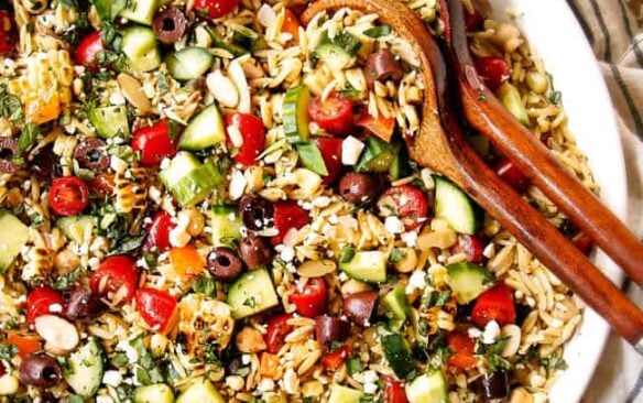 overhead image of orzo pasta salad in a white bowl with wooden spoons.