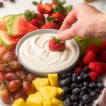close up of a strawberry being dipped into cream cheese fruit dip with fruit all around.