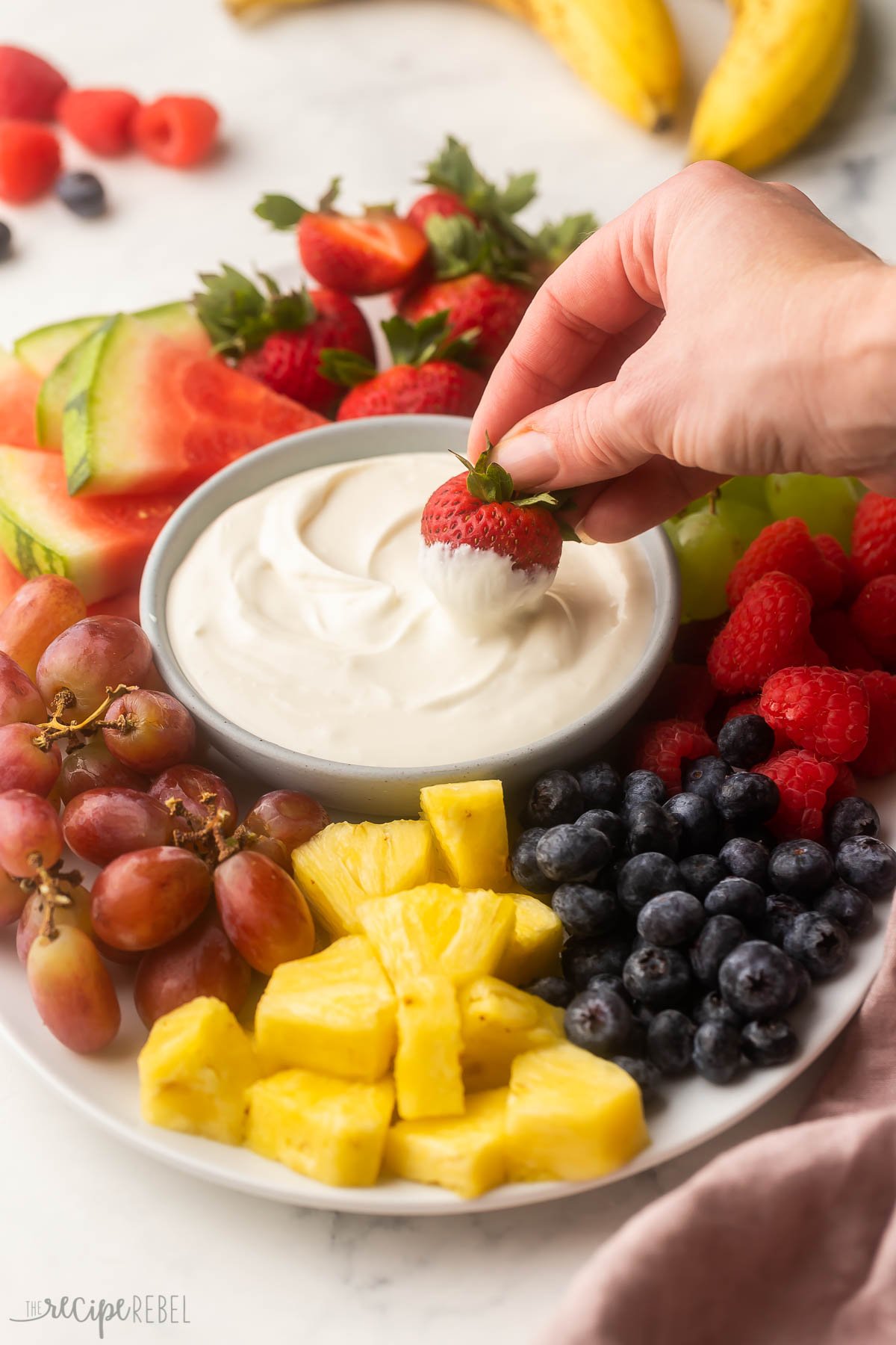 a strawberry being dipped in cream cheese fruit dip amongst a platter of fruit.