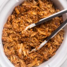 white crockpot filled with pulled pork and steel tongs.