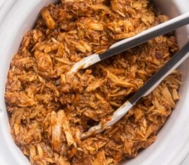 white crockpot filled with pulled pork and steel tongs.