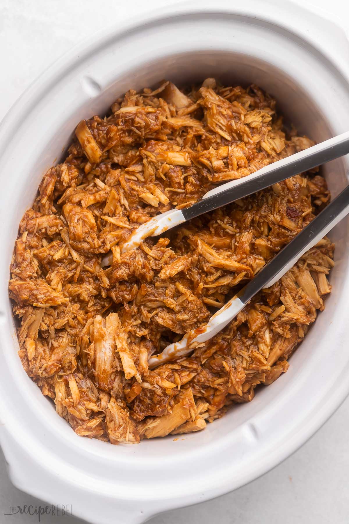 white crockpot filled with pulled pork and steel tongs.