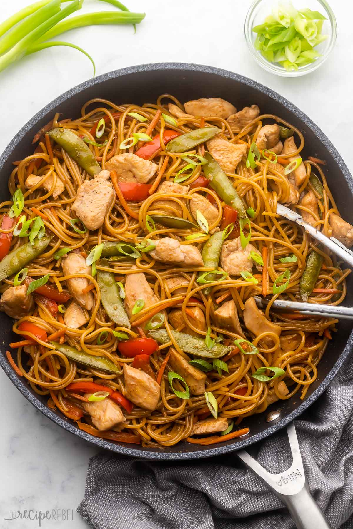 a large black pan filled with teriyaki chicken and noodles on a grey surface with green onions beside.