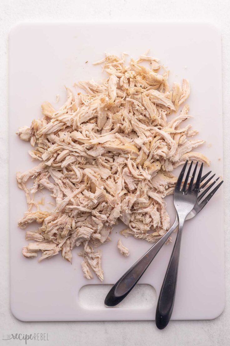 shredded chicken and two forks on a white cutting board.