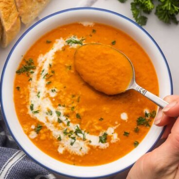 a hand spooning roasted red pepper soup out of white bowl.