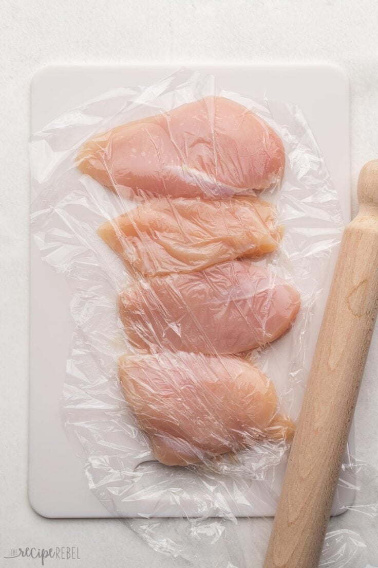 four chicken breasts on cutting board with plastic wrap on top and rolling pin beside.