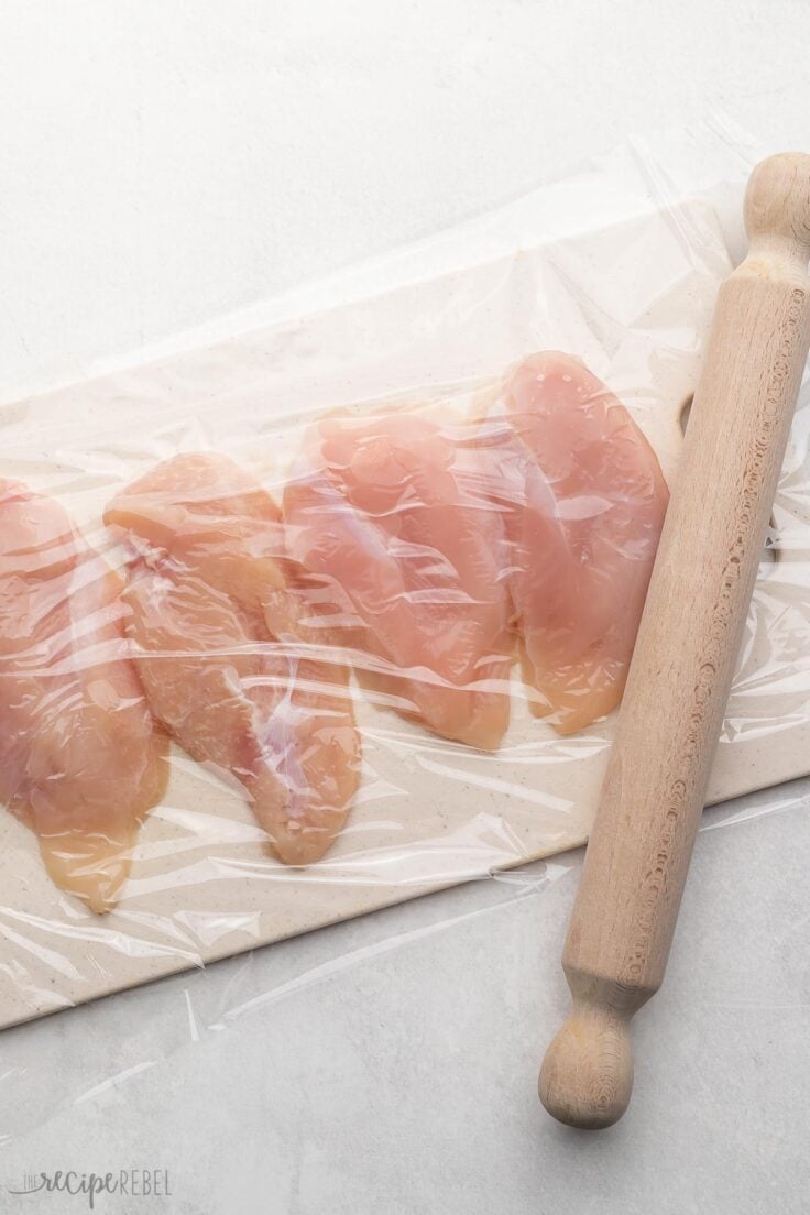 chicken breast on cutting board covered in plastic wrap with rolling pin beside.