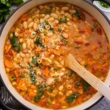 close up view of a large pot of pasta fagioli with a wooden ladle in it and parmesan sprinkled on top.
