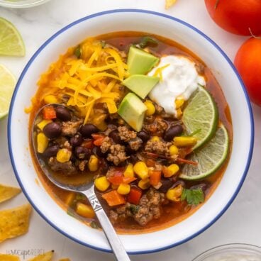 close up view of a white bowl filled with taco soup and topped with avocado, lime, sour cream and cheese.