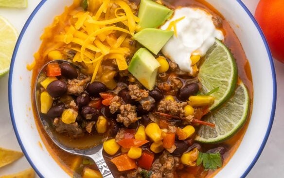 close up view of a white bowl filled with taco soup and topped with avocado, lime, sour cream and cheese.