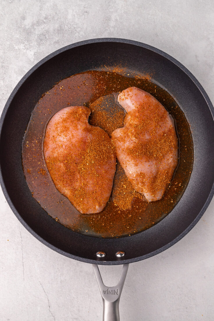 black frying pan with two seasoned chicken breasts inside.