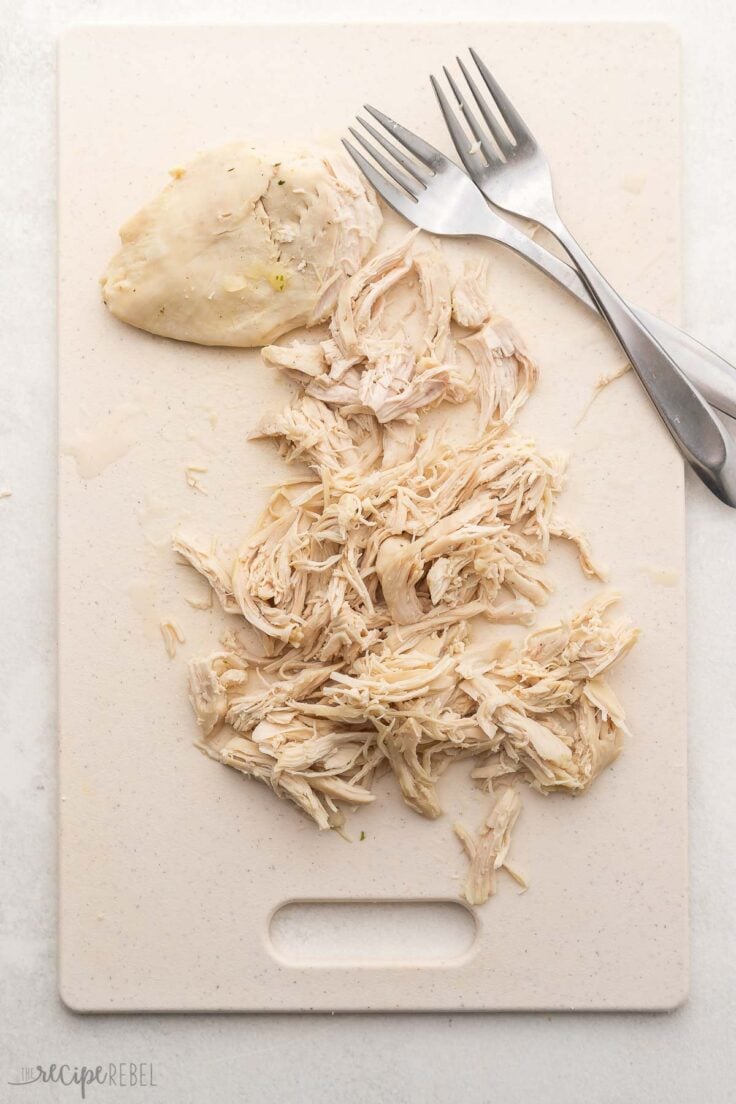 white cutting board with partially shredded chicken breast and two forks.