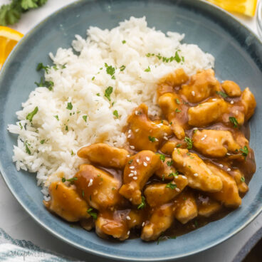 orange chicken and white rice in a bowl topped with chopped parsley.