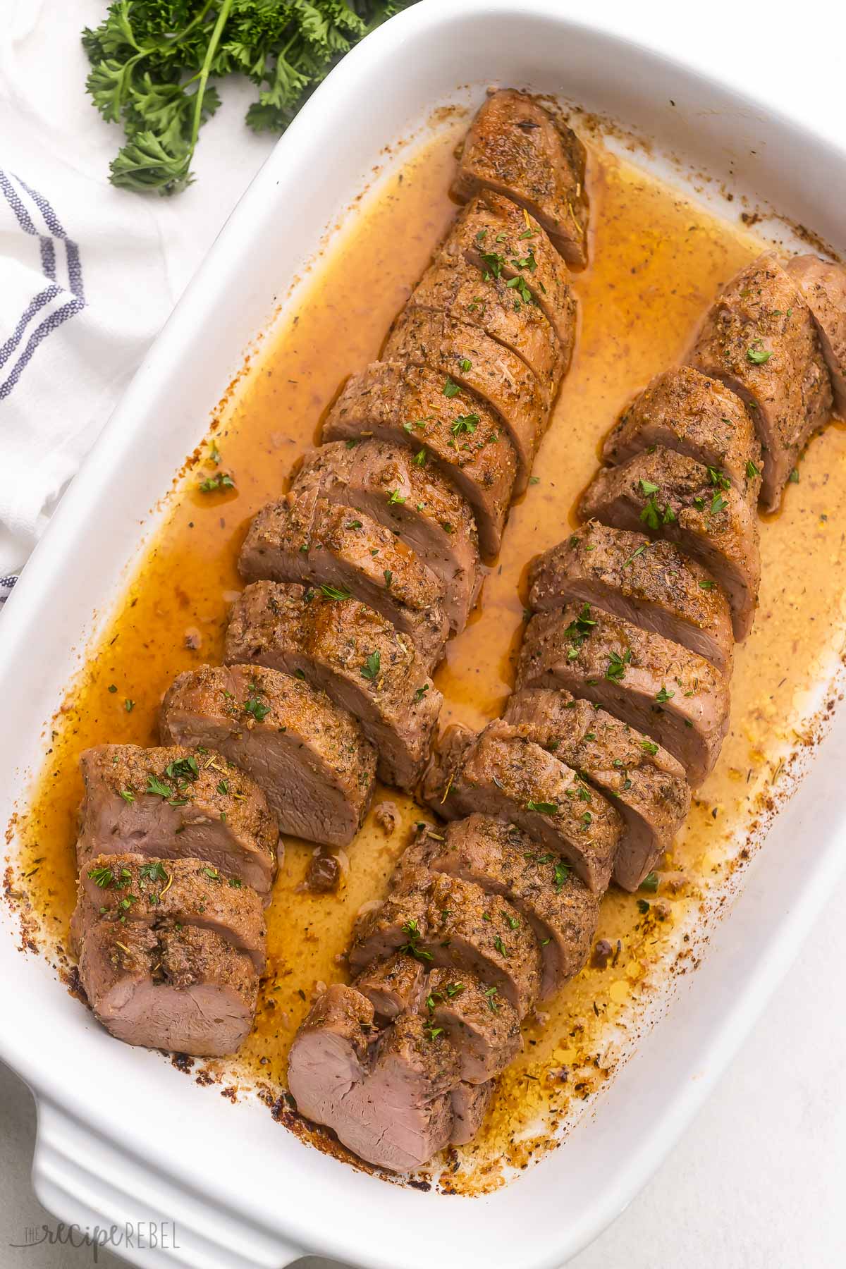 overhead view of sliced pork tenderloin lying in a white baking dish with parsley beside.
