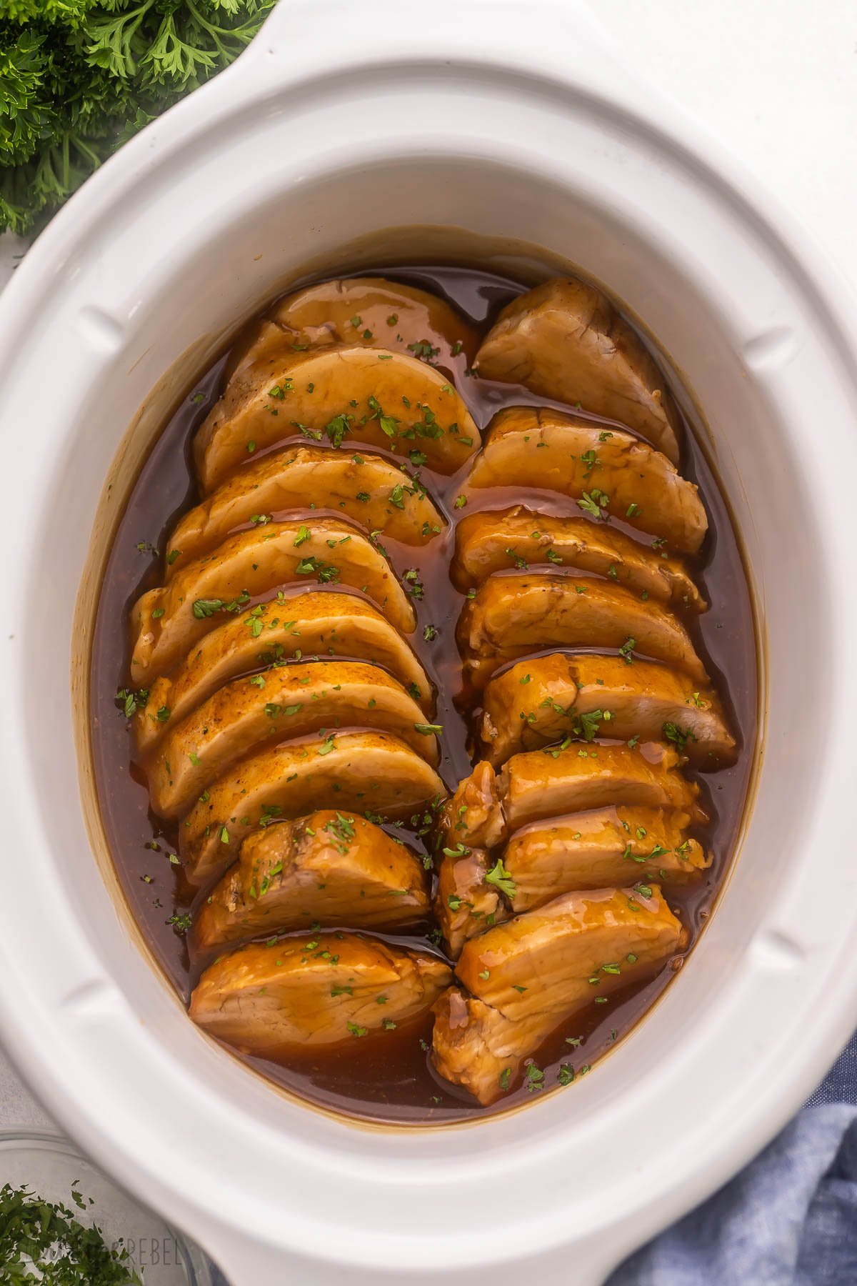 overhead view of sliced pork tenderloin in white slow cooker topped with chopped parsley.