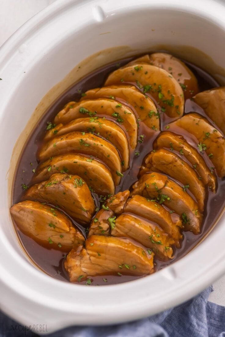 sliced pork tenderloin in white slow cooker topped with chopped parsley.