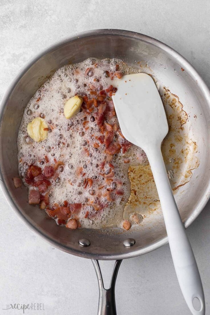 steel frying pan with cooked bacon, garlic, and a spatula.