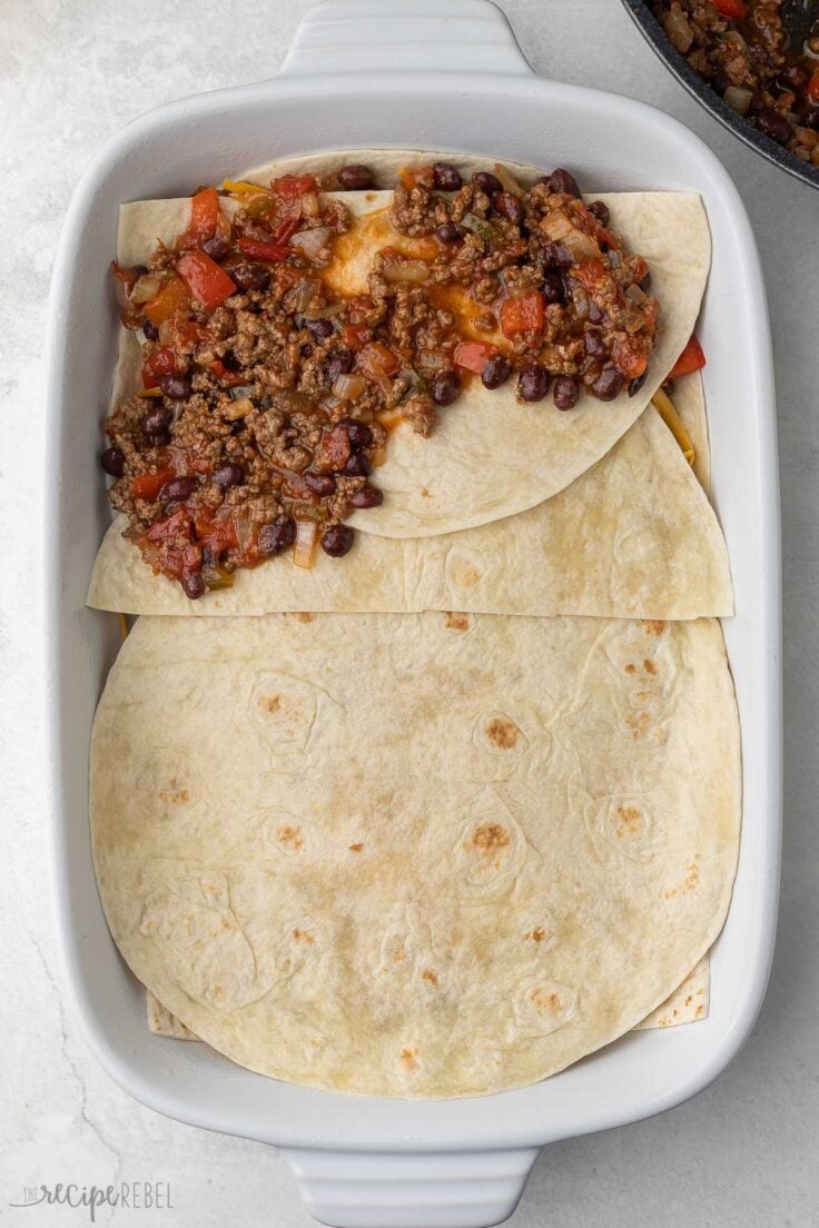 taco meat and seasoning partially spread across tortillas in baking dish.