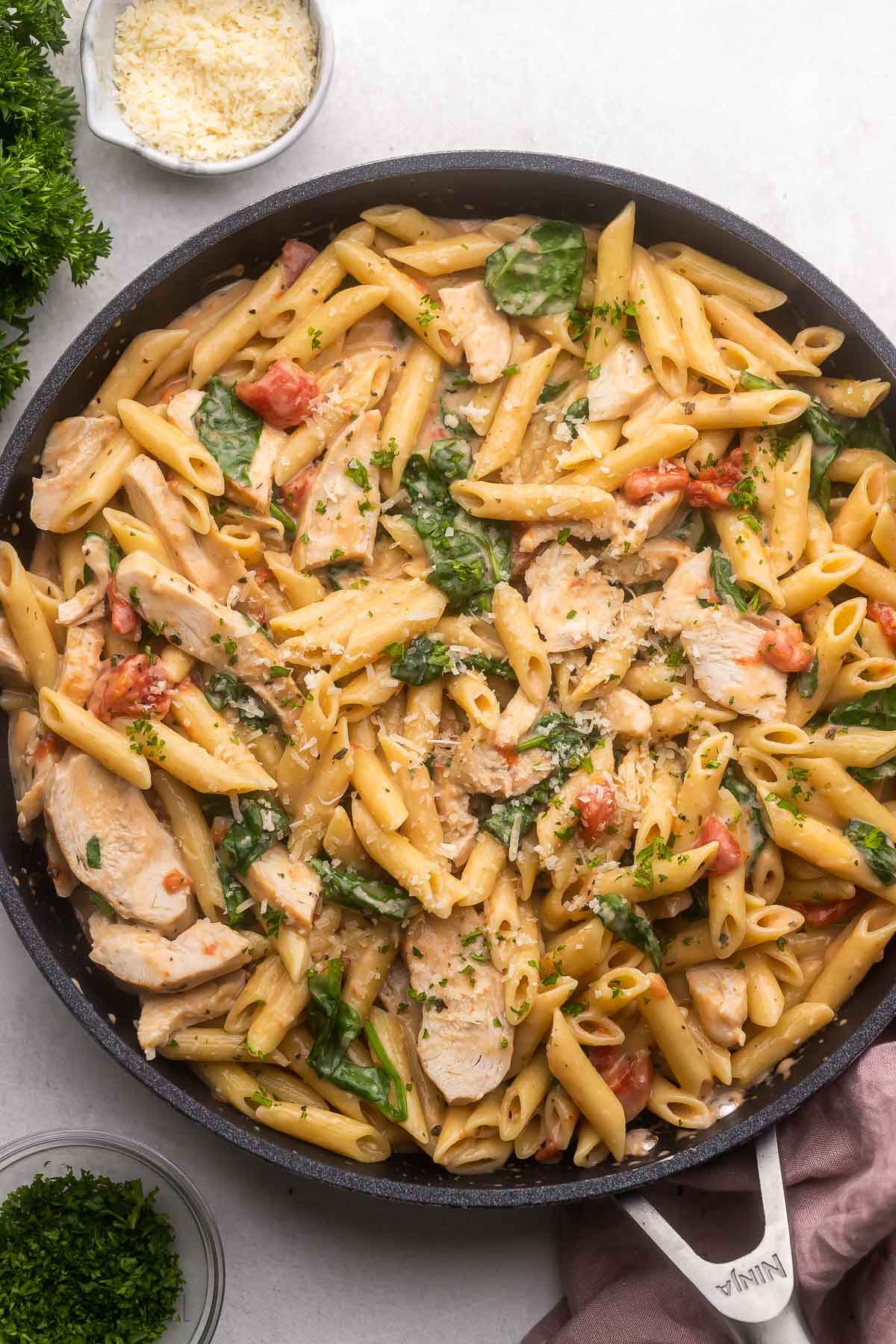 overhead view of a black pan filled with chicken penne pasta with parmesan and parsley beside.