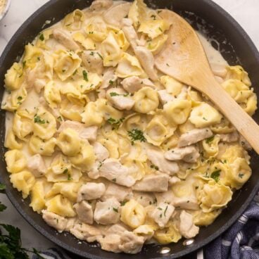 large black pan filled with chicken tortellini alfredo and wooden ladle.