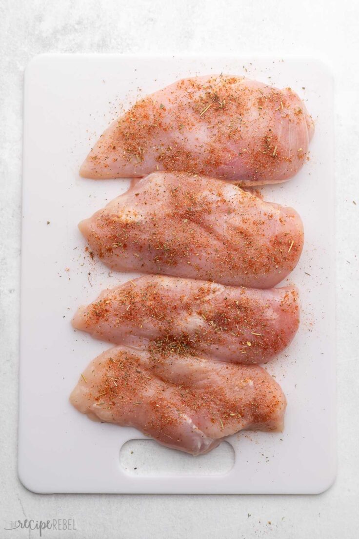 four seasoned chicken breasts on a white cutting board.