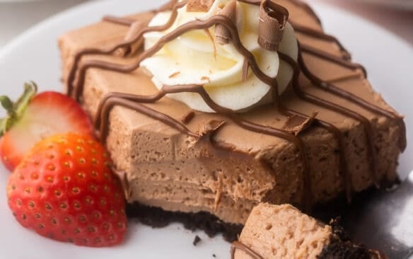 white plate with a nutella cheesecake bar drizzled with chocolate and strawberries beside.