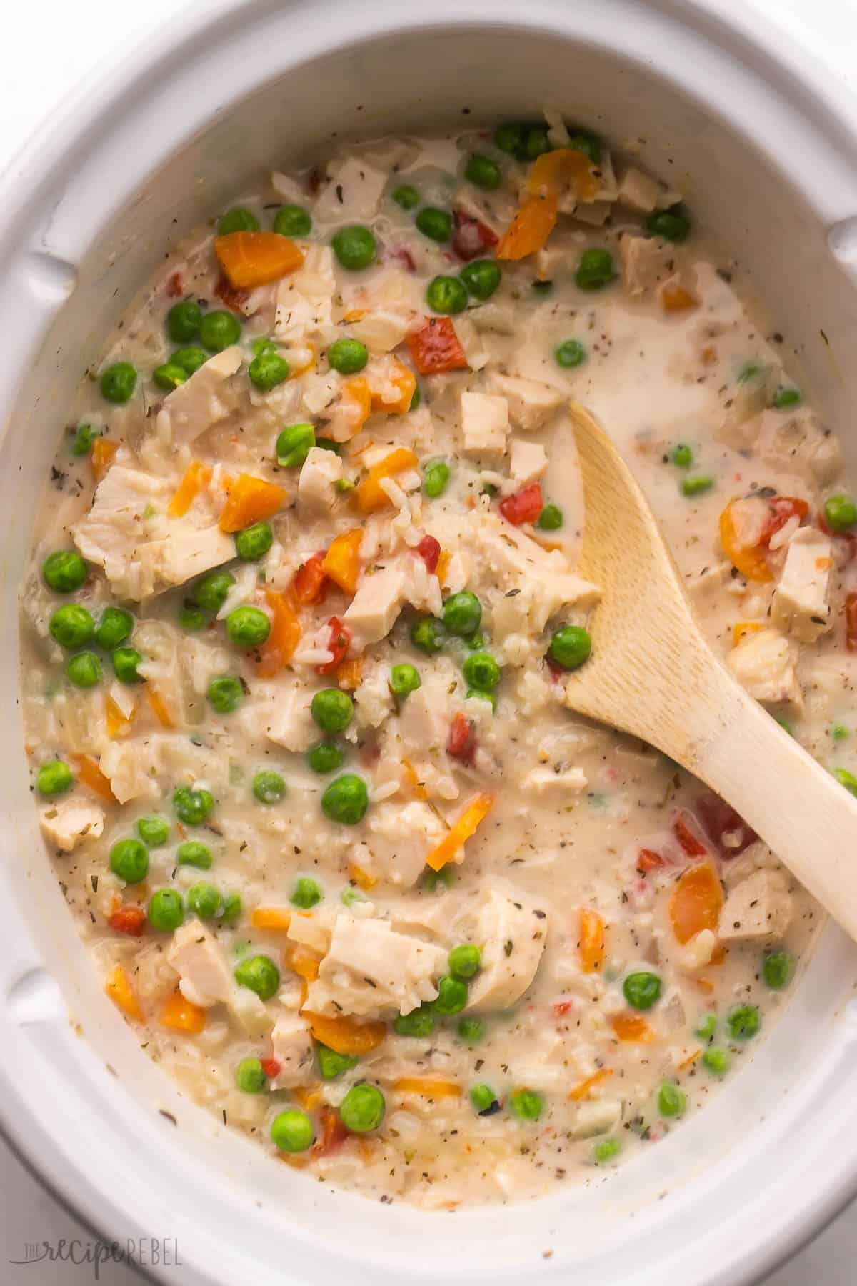 Slow Cooker Chicken and Rice features bites of tender chicken breasts, a medley of veggies, a creamy, cheesy sauce and perfectly cooked white rice. It's an easy dinner recipe that the whole family loves! #slowcooker #crockpot #chicken #dinner #recipe | dinner recipes | easy recipes | slow cooker recipes | crockpot dinner | crockpot chicken recipe | chicken and rice | chicken breast | family dinner a wooden ladle inside a slow cooker filled with chicken and rice.