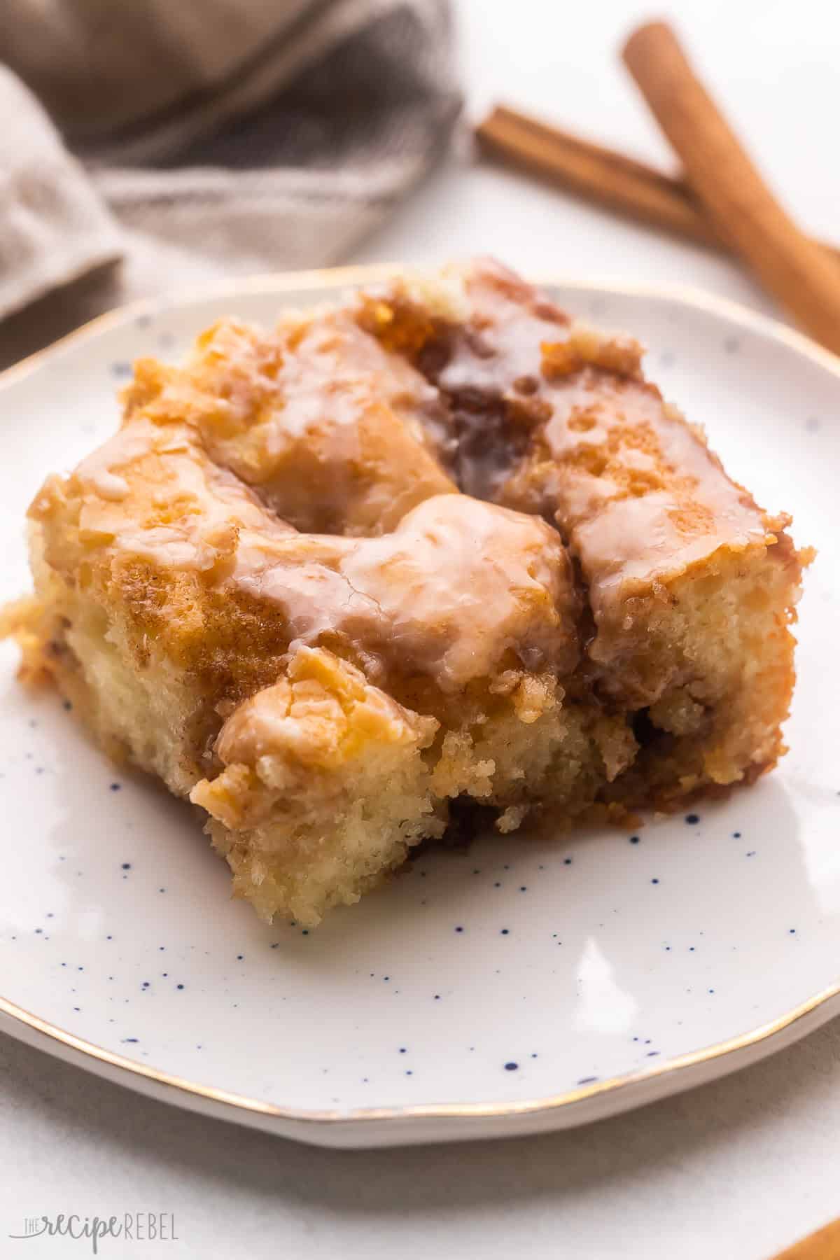a piece of cinnamon roll cake on a white plate with cinnamon sticks behind it.