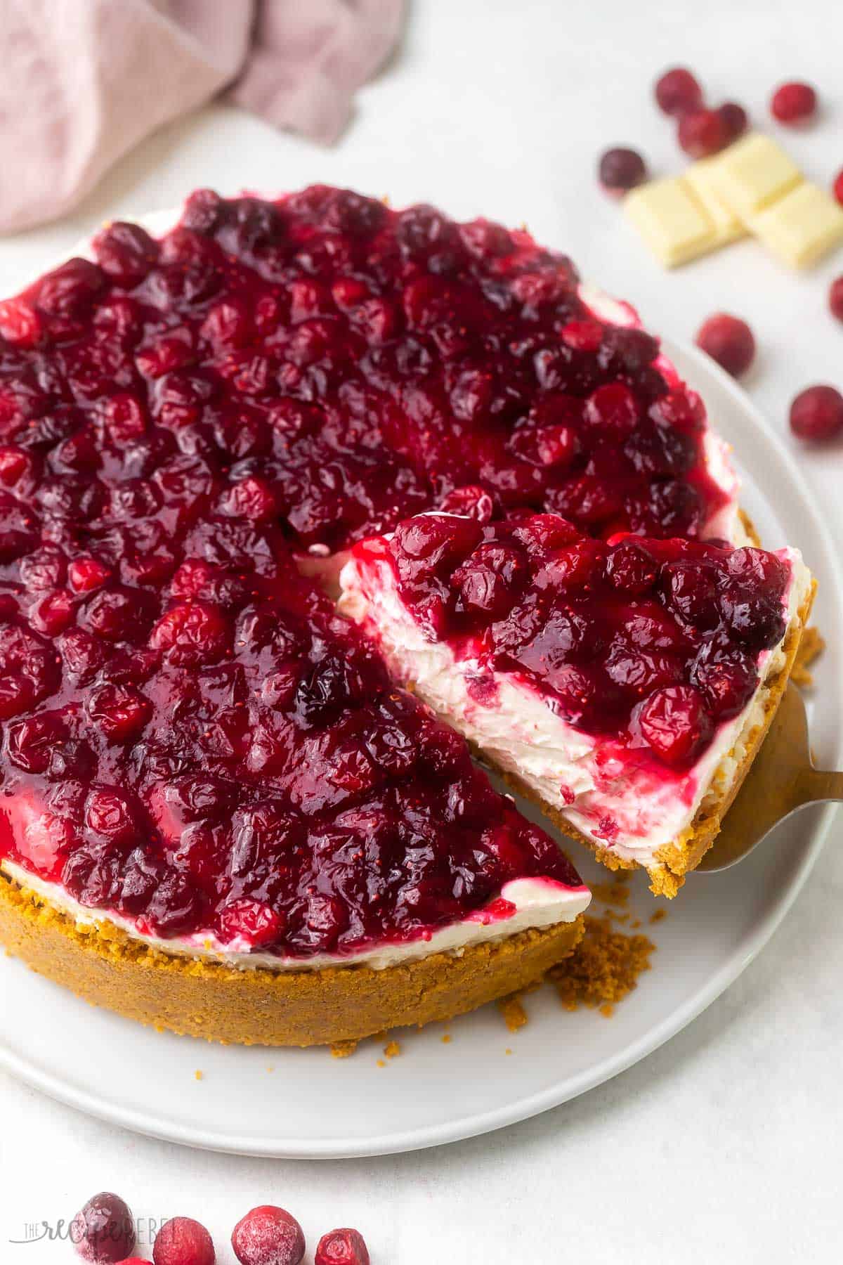 This No Bake White Chocolate Cranberry Cheesecake has the perfect balance of sweet and tart flavors! It's a smooth, creamy, luscious cheesecake that doesn't require a water bath or a hot oven. I think my favorite part might just be the vibrant cranberry sauce that's spread all across the top! #cheesecake #nobake #dessert #recipe | no bake dessert | dessert recipes | cheesecake recipes | cranberry dessert | christmas dessert | christmas cheesecake | thanksgiving dessert | cranberry sauce cranberry white chocolate cheesecake on a large white plate with cranberries and chocolate beside.