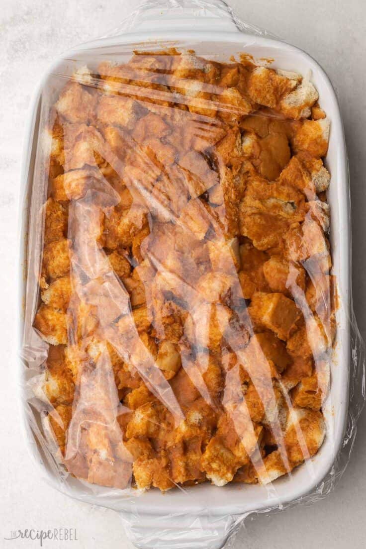 unbaked pumpkin french toast casserole in white dish covered in plastic wrap.