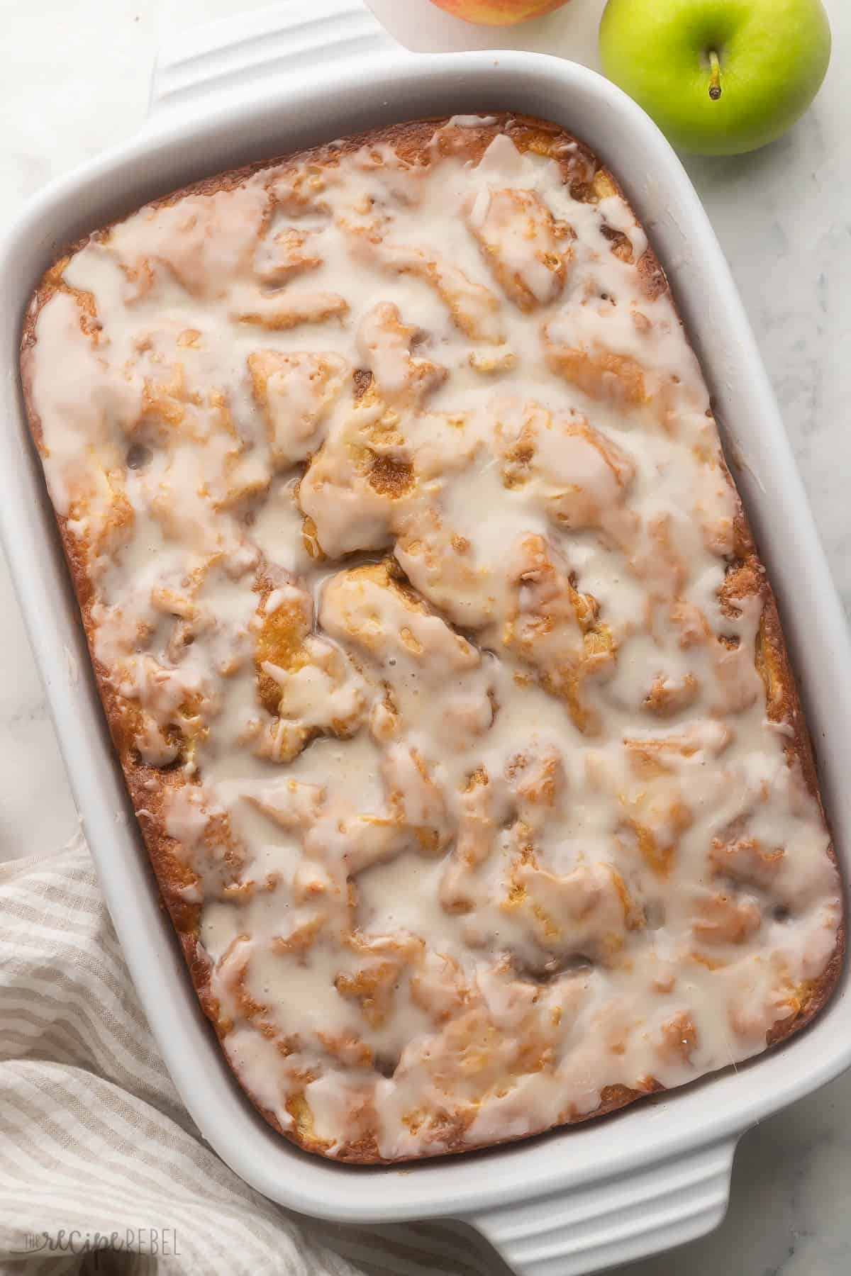 This Apple Fritter Cake recipe has all the flavors of an apple fritter but in cake form! A moist cake made with Greek yogurt, oil, tender apples, and warm spices, swirled with cinnamon sugar and topped with a sweet glaze for the ultimate fall treat. #apple #dessert #cake | apple recipe | baking recipe | apple cake | bake from scratch | apple desserts | fall desserts | fall baking | christmas dessert | thanksgiving large white pan of apple fritter cake with apples beside.