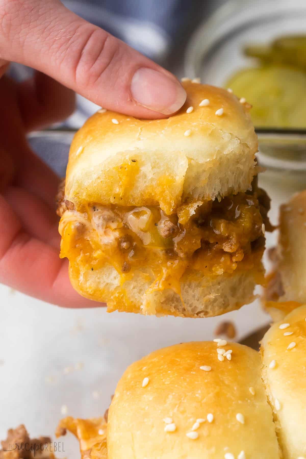 These easy Cheeseburger Sliders take less than 30 minutes to make and are perfect for parties, game day, or any time you need a crowd-pleasing appetizer! Fluffy Hawaiian rolls are layered with melty cheddar cheese, lean ground beef cooked in a special sauce, and topped with melted butter and sesame seeds. #appetizer #groundbeef #recipe | ground beef recipes | dinner ideas | cheeseburgers | game day food | party food a hand holding a cheeseburger slider with pickles in the background.